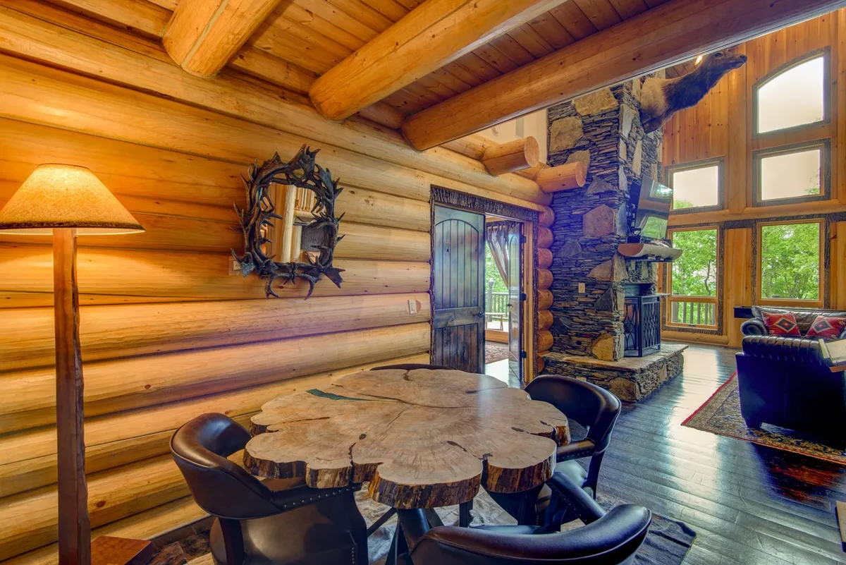 A welcoming gathering space with log walls, a striking stone fireplace, a unique wooden table, large windows, and plush seating. The setting is perfect for group meals, game nights, or relaxing by the fire while taking in scenic views.