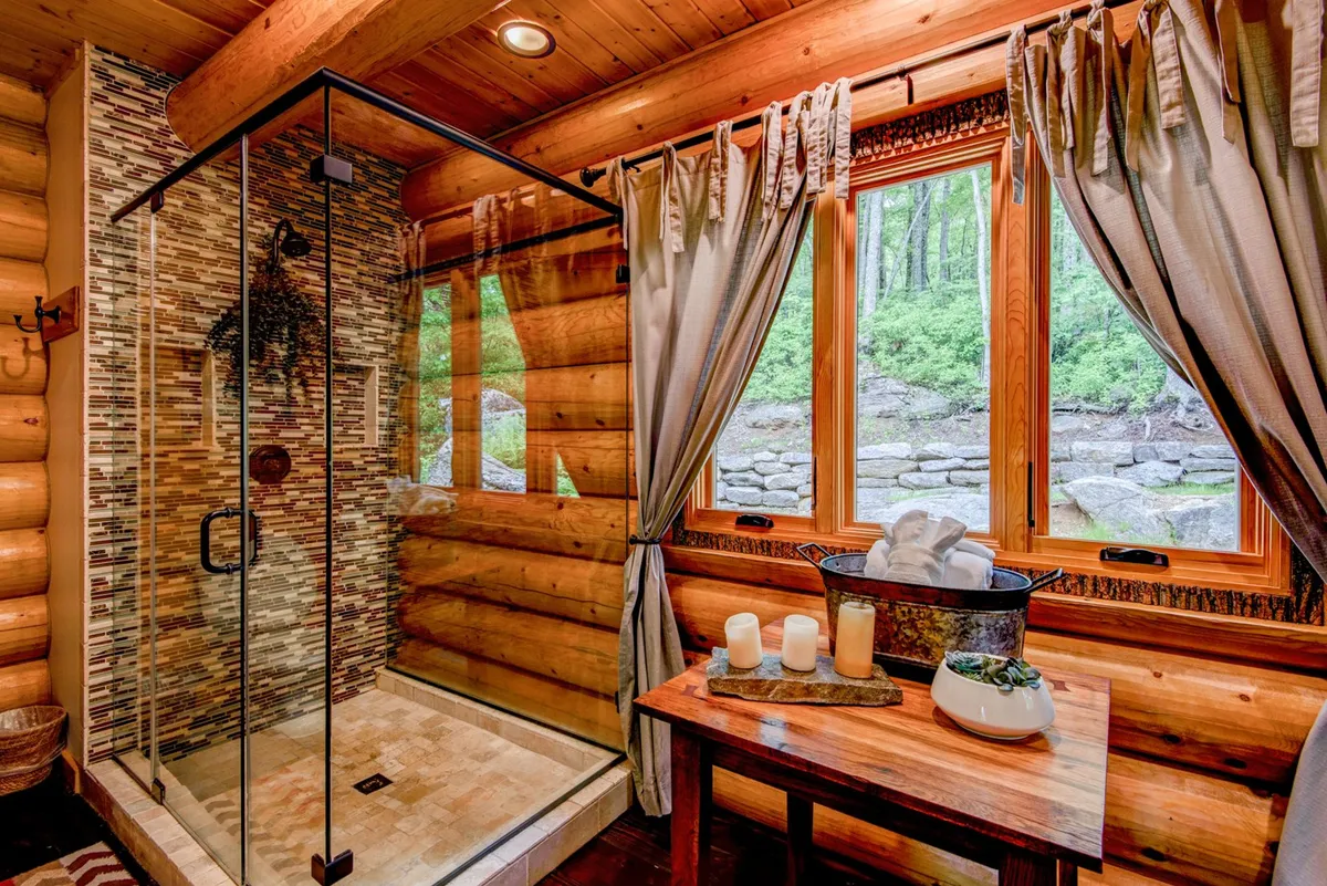 A beautifully appointed bathroom with a glass walk-in shower, natural wood elements, a large window offering outdoor views, and calming decorative touches like candles and towels. Morning light pours into the space, creating a soothing and luxurious atmosphere.