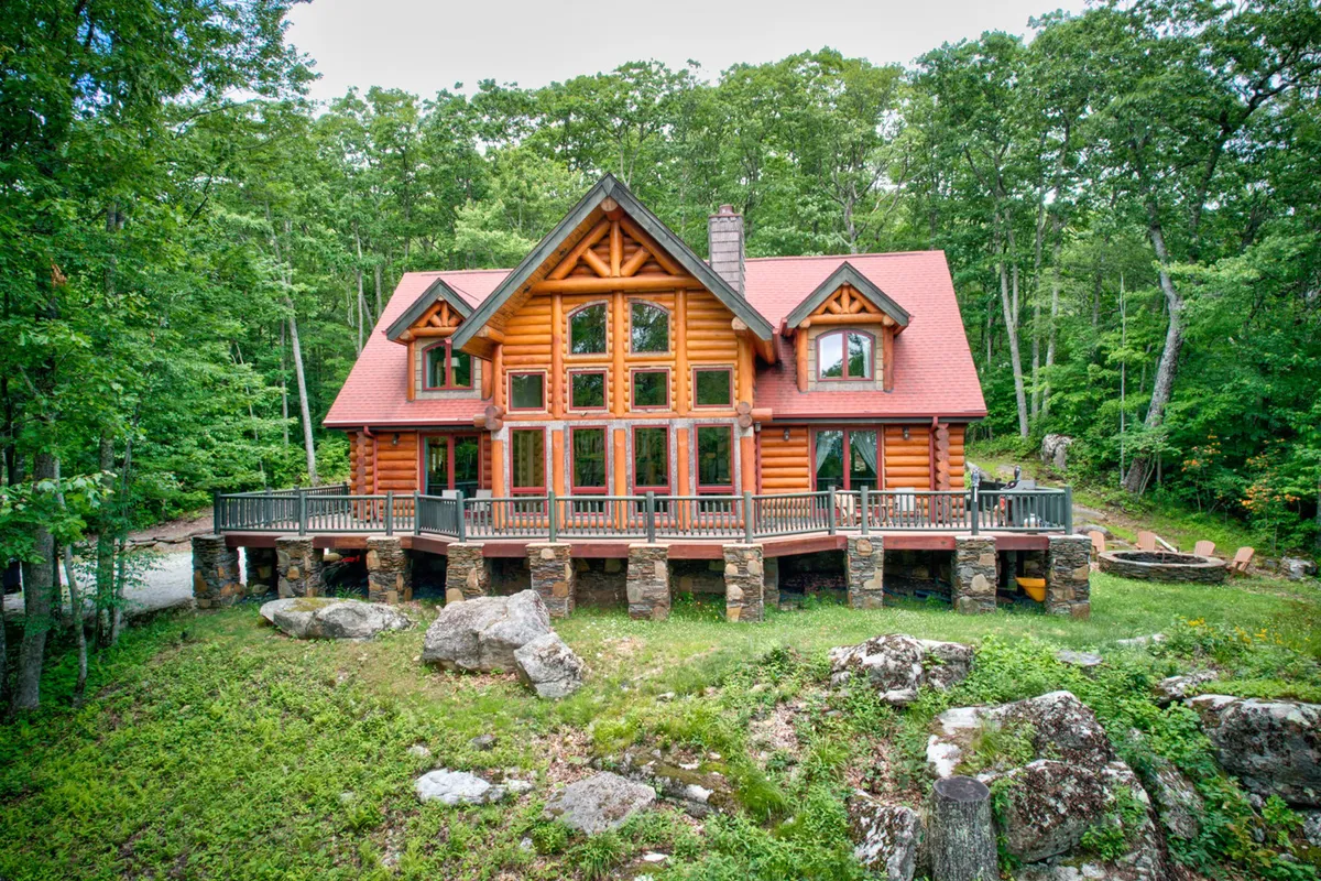 This exterior shot beautifully captures the log cabin, large windows, expansive deck area, and surrounding lush greenery. A fire pit is visible in the foreground, suggesting an inviting space for group gatherings and cozy evenings under the stars. The setting sun bathes the scene in a warm glow, adding to the atmosphere of camaraderie and relaxation.
