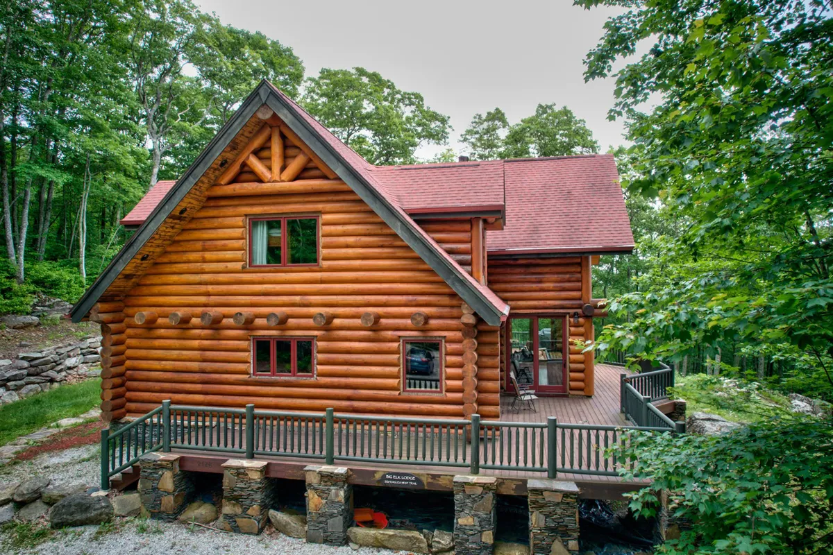 A rustic log cabin, surrounded by trees, with a spacious deck area and a sturdy stone foundation. The deck is an inviting space for gatherings, while the woodsy setting emphasizes proximity to nature and trail access. The scene suggests outdoor relaxation and easy transition from the lodge to the wilderness.