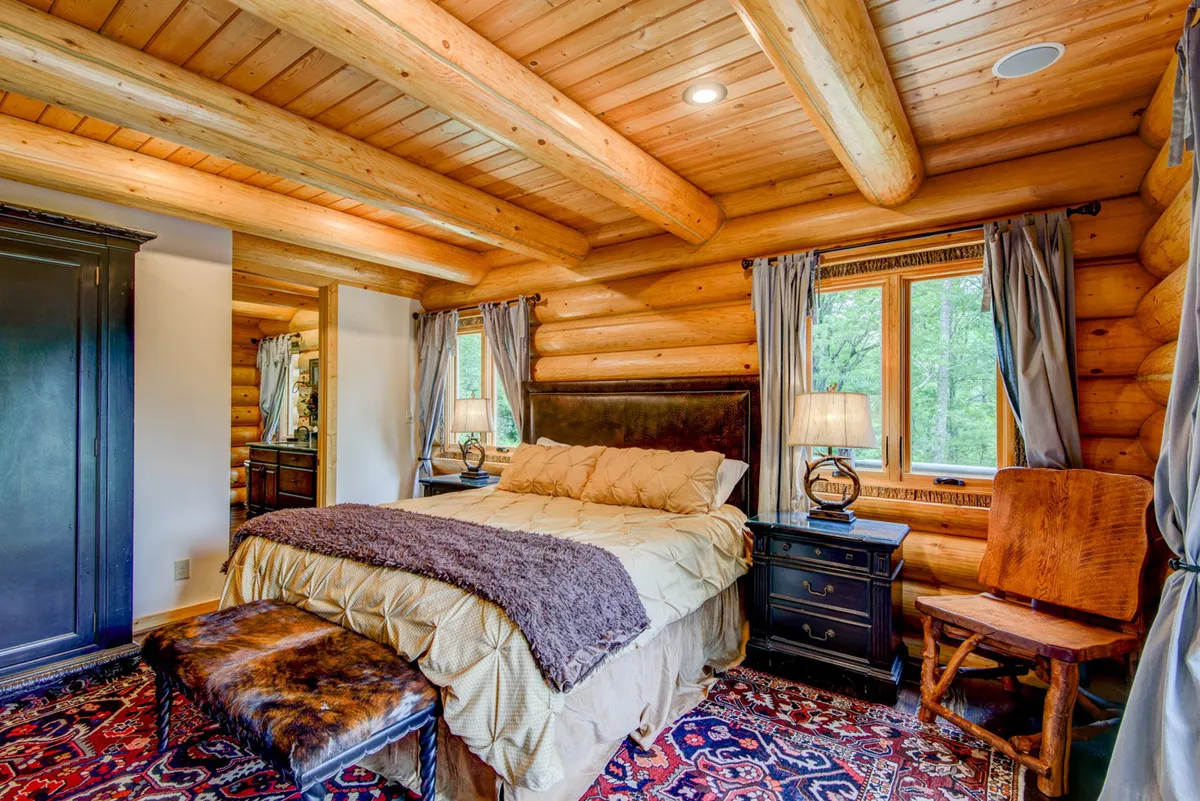 A cozy and luxurious bedroom featuring log walls, a plush king-size bed with inviting bedding, nightstands with softly glowing lamps, a decorative rug, and a large window letting in warm sunlight. The space radiates tranquility and warmth, perfect for relaxation.