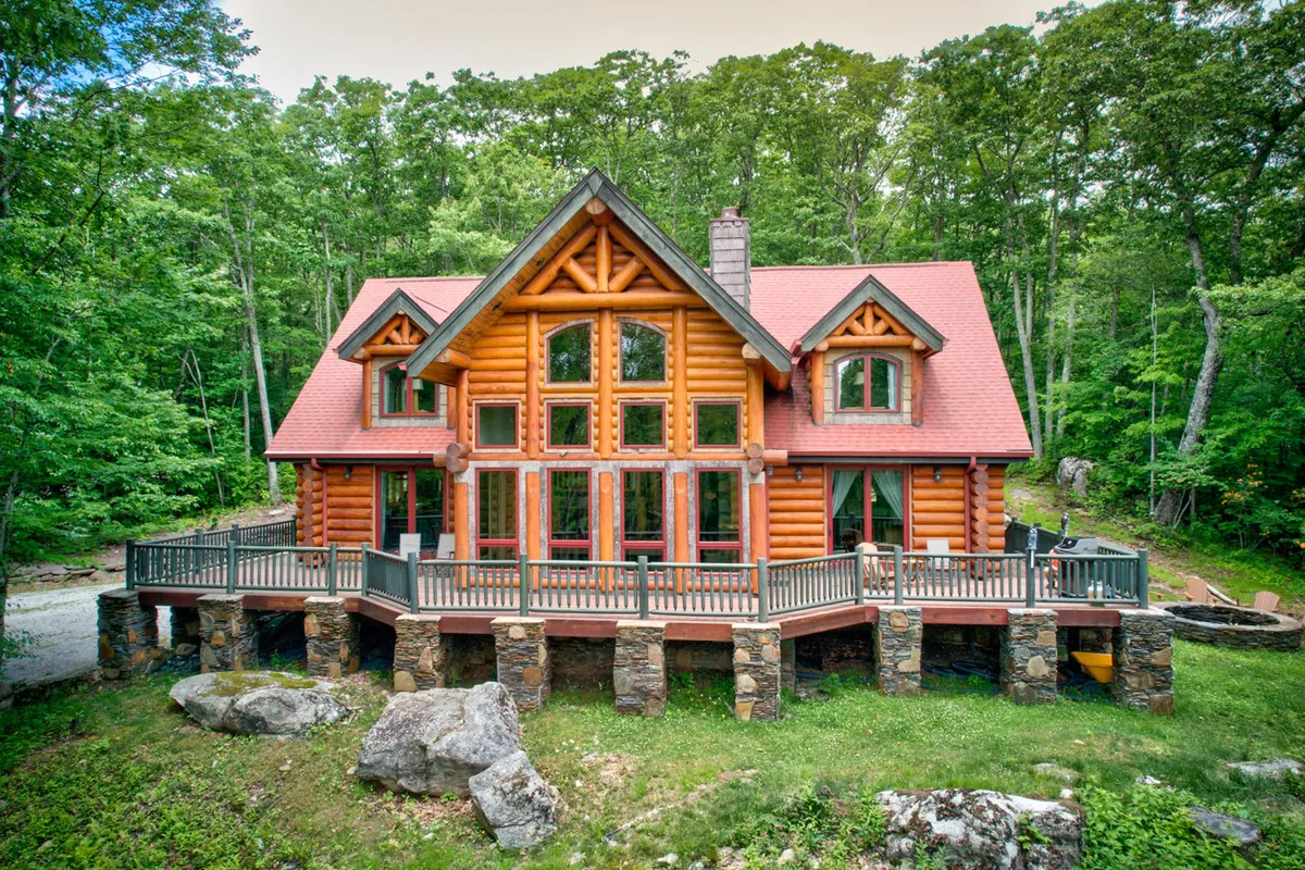 The hero image showcases a grand log cabin set amidst the wooded surroundings of Banner Elk. The cabin features impressive solid log construction, expansive windows that capture natural light, a sturdy stone foundation, and a spacious deck perfect for relaxing or socializing. The inviting outdoor area hints at amenities like a fire pit for evening gatherings and breathtaking views of the surrounding forest.
