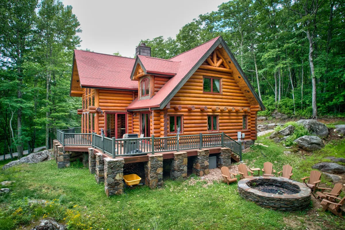 A cozy log cabin with a wooden deck and a prominent stone fire pit set among tall trees. There is comfortable seating around the fire pit, and the deck invites relaxation with its tranquil forest backdrop.