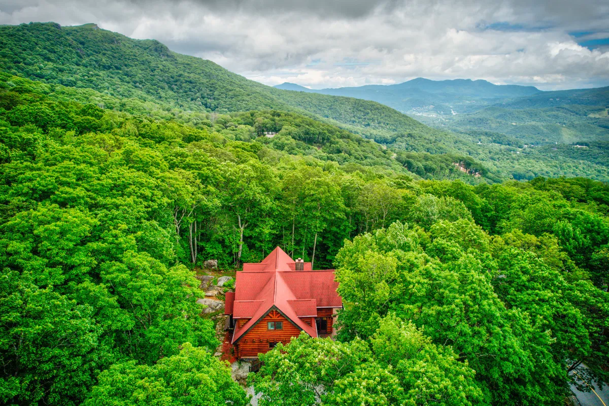 A red-roofed cabin nestled among lush green trees with distant mountain views, set against a serene backdrop. The porch and yard hint at spaces for bonfires, relaxing, and prepping for outdoor activities. The setting is tranquil yet ripe for exploration.