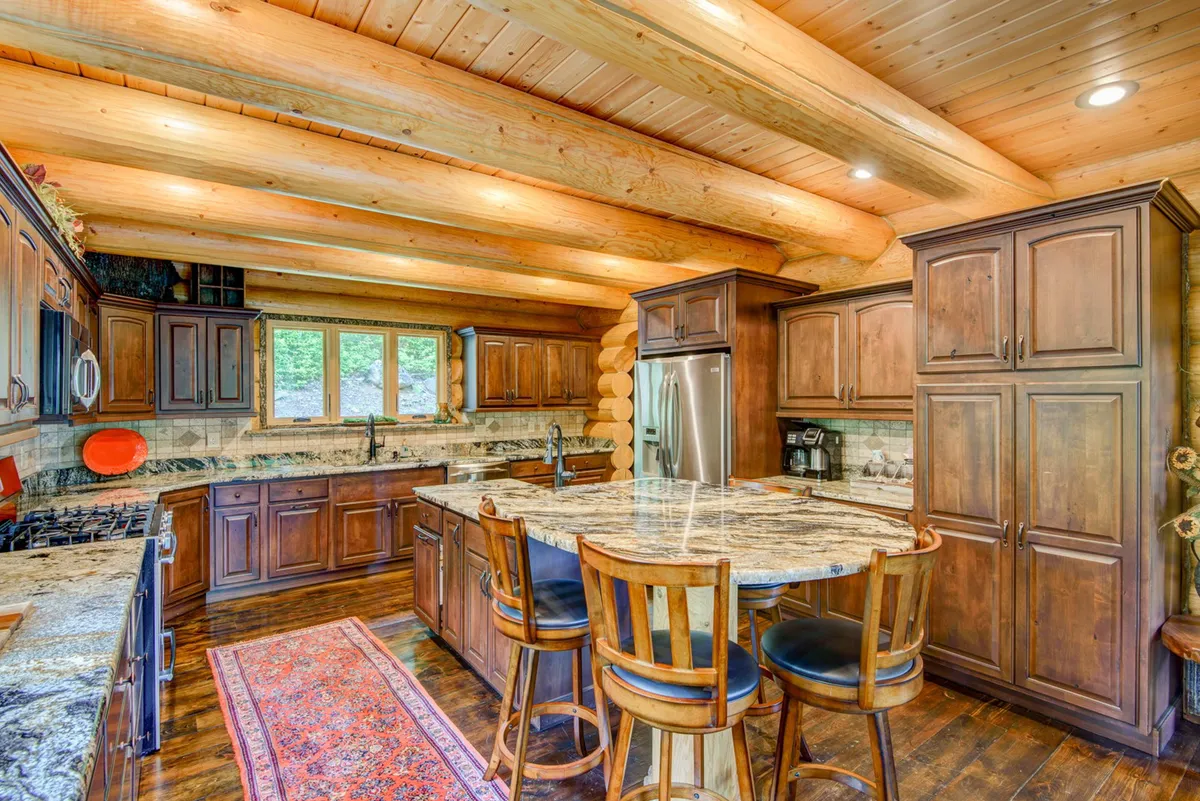 A bright kitchen scene with granite countertops, wooden cabinets, stools arranged around the island, and expansive windows flooding the area with sunlight. The setup encourages casual dining and conversation, with a welcoming ambiance.
