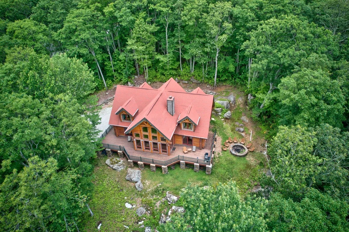A wide exterior shot featuring the house, a fire pit area, and the surrounding trees. The setup is clearly intended for outdoor group gatherings, with seating around the fire pit and open space for relaxation or storytelling under the stars.