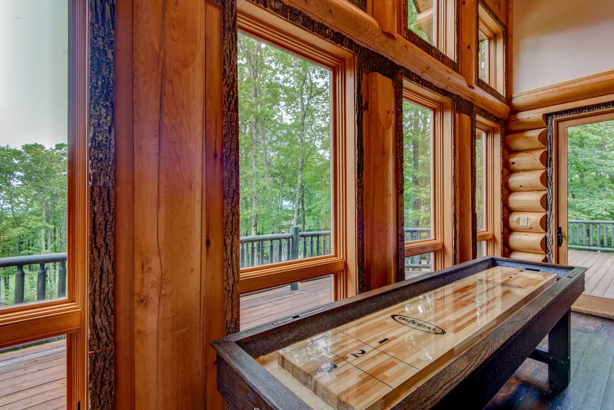 A relaxed lounge area with a shuffleboard table positioned near expansive windows, exposed wooden beams overhead, and a cozy, social atmosphere ideal for friendly games and gatherings. The sunset glow through the windows adds to the inviting mood.