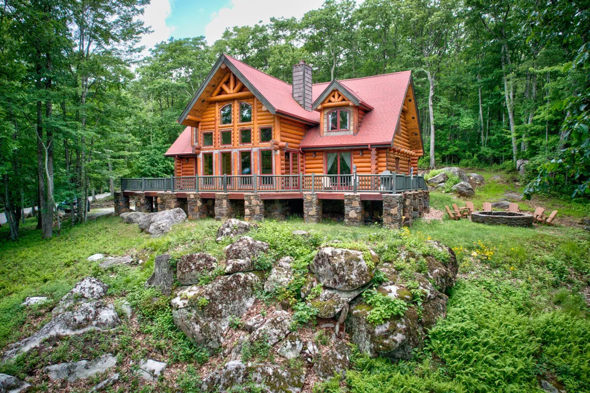 A log cabin nestled in greenery with a dedicated firepit area, outdoor seating, and a large deck. The firepit is set up for gatherings, and the overall scene is tranquil and inviting, perfect for evening stargazing.