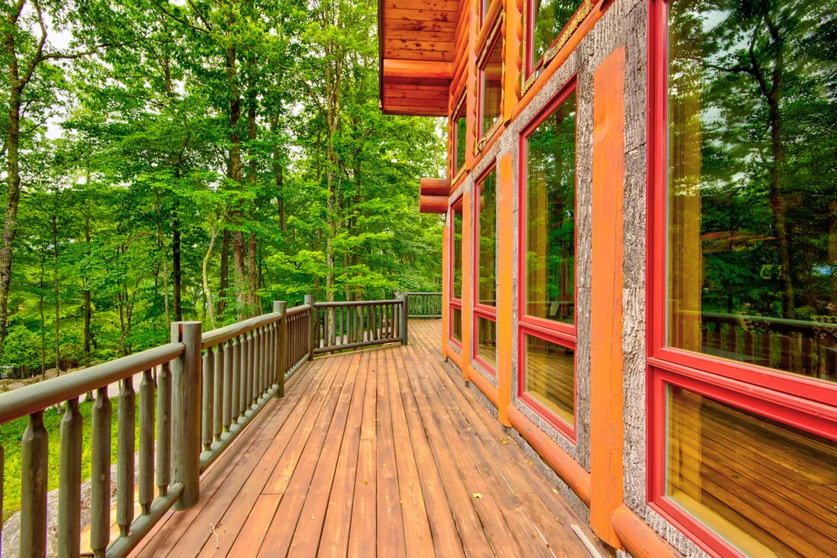 This image shows a spacious wooden deck attached to the lodge, bordered by large windows that look out onto a serene, natural landscape. The deck appears inviting for relaxation or socializing, with plenty of room for outdoor seating. Natural surroundings hint at mountain views and quiet evenings under the stars.