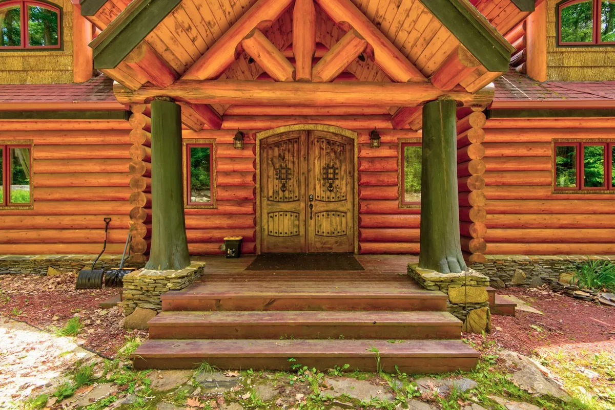 An inviting view of the lodge's ornate wooden doors, log cabin structure, and stone steps, with people gathering on the porch. The sunset in the background and hints of a fire pit create an atmosphere ideal for welcoming groups and enjoying the outdoors together.