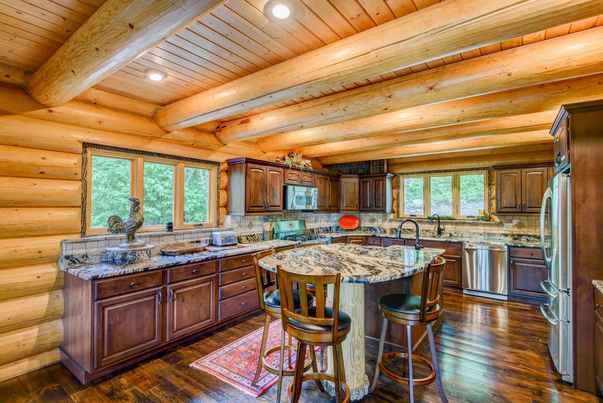 A spacious kitchen with a granite countertop, wooden cabinets, log walls, a kitchen island, and large windows that let in natural light. The space is warm and inviting, with plenty of room for meal preparation and family gatherings.