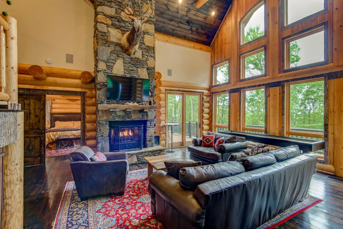 A spacious great room featuring a large stone fireplace, comfortable leather sofas, a rug with intricate design, antler decoration, and floor-to-ceiling windows showcasing beautiful mountain vistas. The warm lighting and arrangement invite groups to gather and relax together.