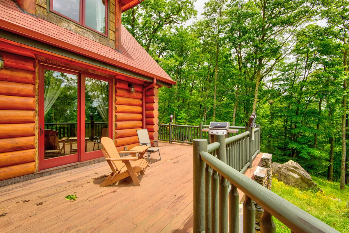 A log cabin with a spacious deck surrounded by lush greenery, featuring comfortable outdoor seating. The scene emphasizes the integration of the deck with the natural environment, ideal for morning coffee or sunset views.